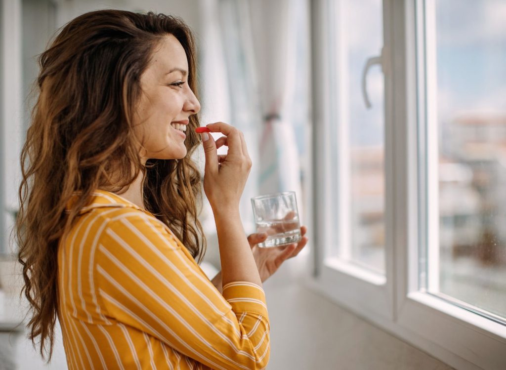 A woman smiling holding a supplement to her mouth with one hand and a glass of water in the other.