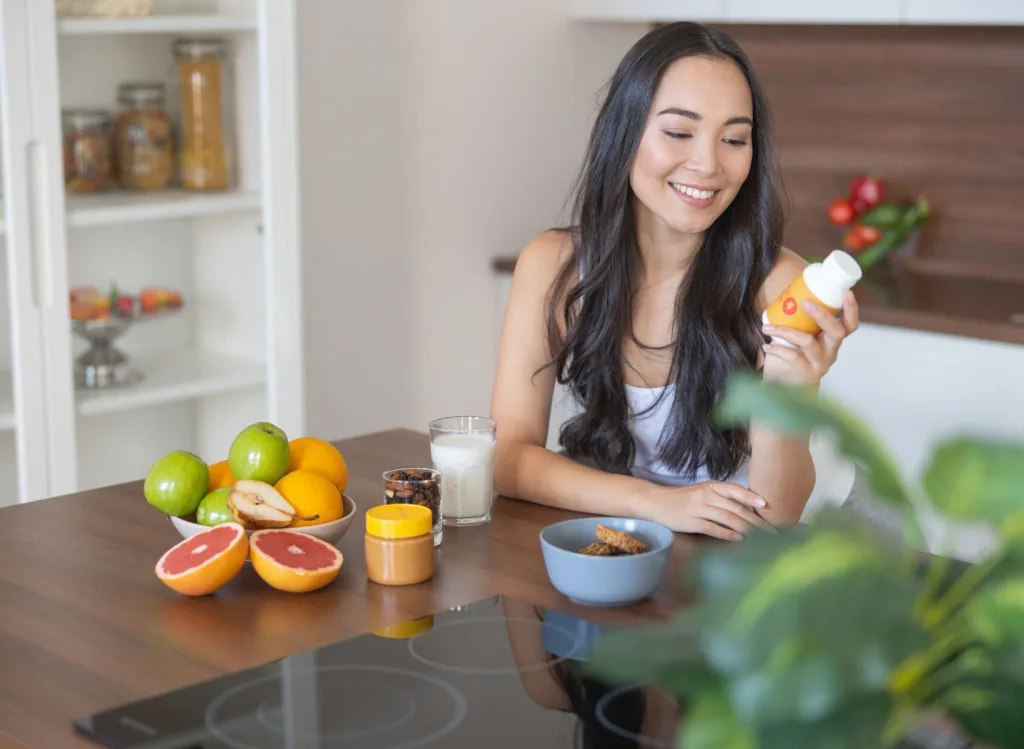 Woman in a kitchen area looking at a bottle of supplements surrounded by various fruit.