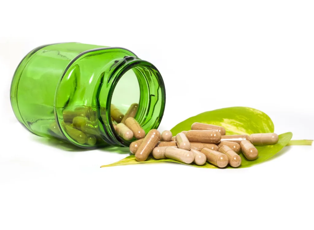 A green transparent glass bottle on its side with filled capsules spilling out onto a leaf.