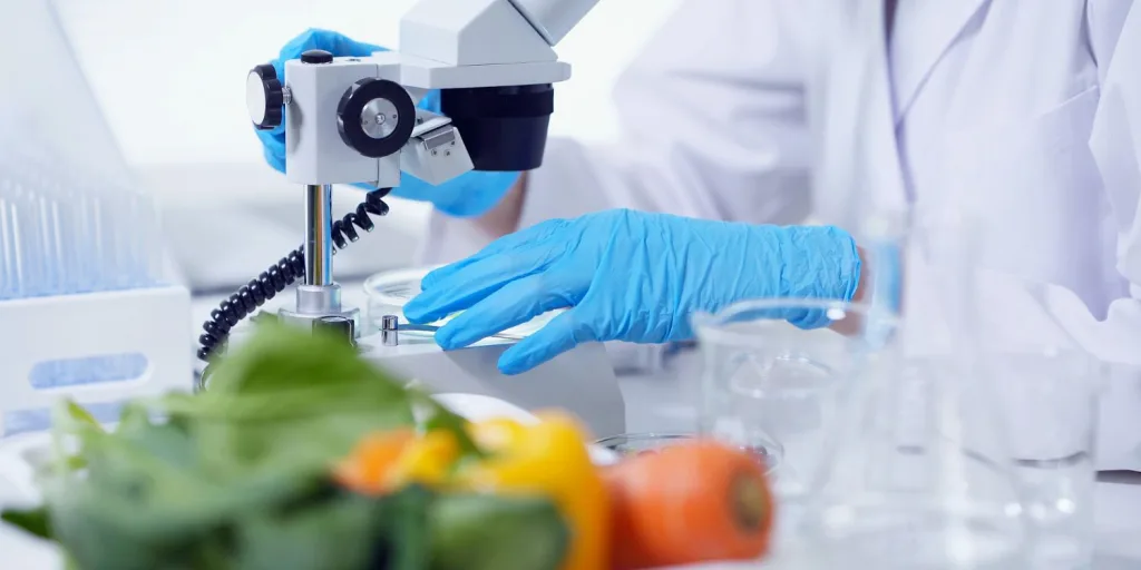 Person in lab PPE handling a microscope with vegetables in the foreground.