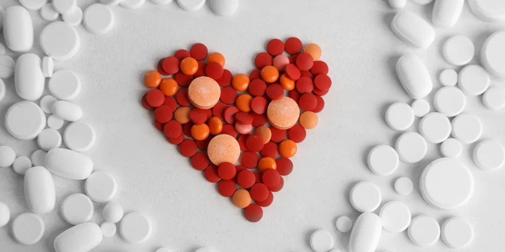 Red and orange different sized round tablets compiled in a shape of a heart, surrounding by varying sizes and shapes of white tablets.