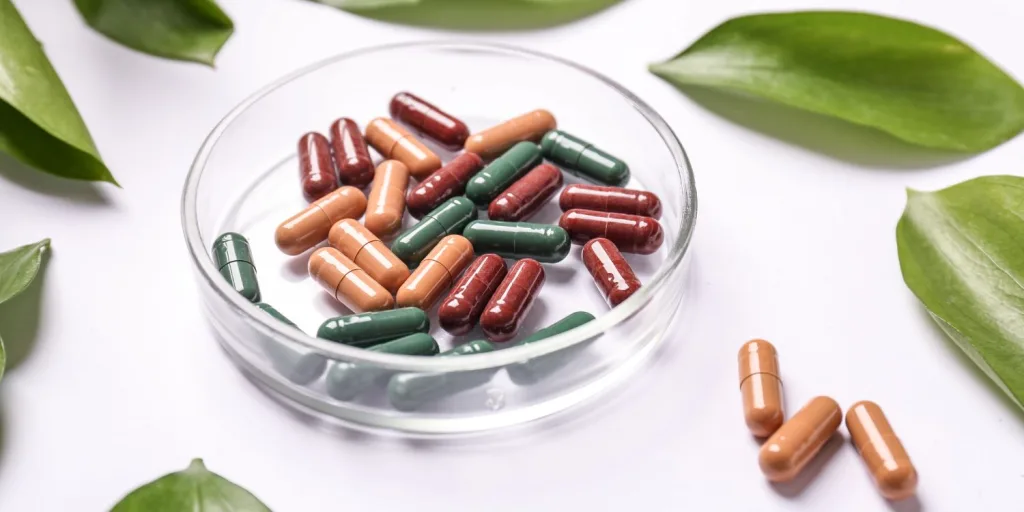 Assorted dark red, green, and orange capsules in a petri dish, surrounded by green plant leaves.