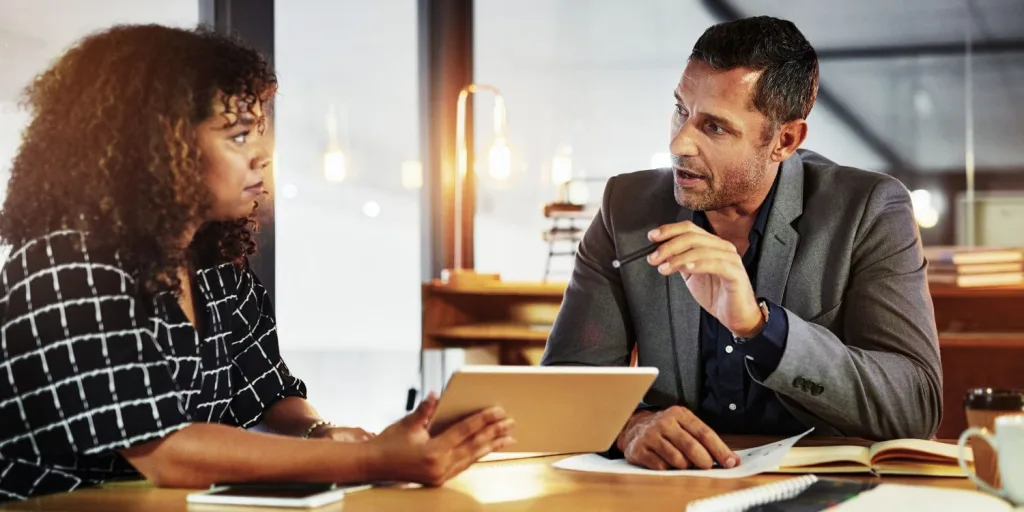 A woman and a man talking while looking at a tablet in a professional setting.