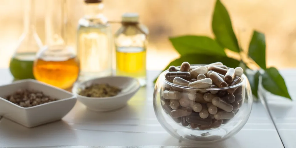 An assortment of filled capsules in a small glass bowl. There are liquid ingredients as well as powder ingredients in the background.