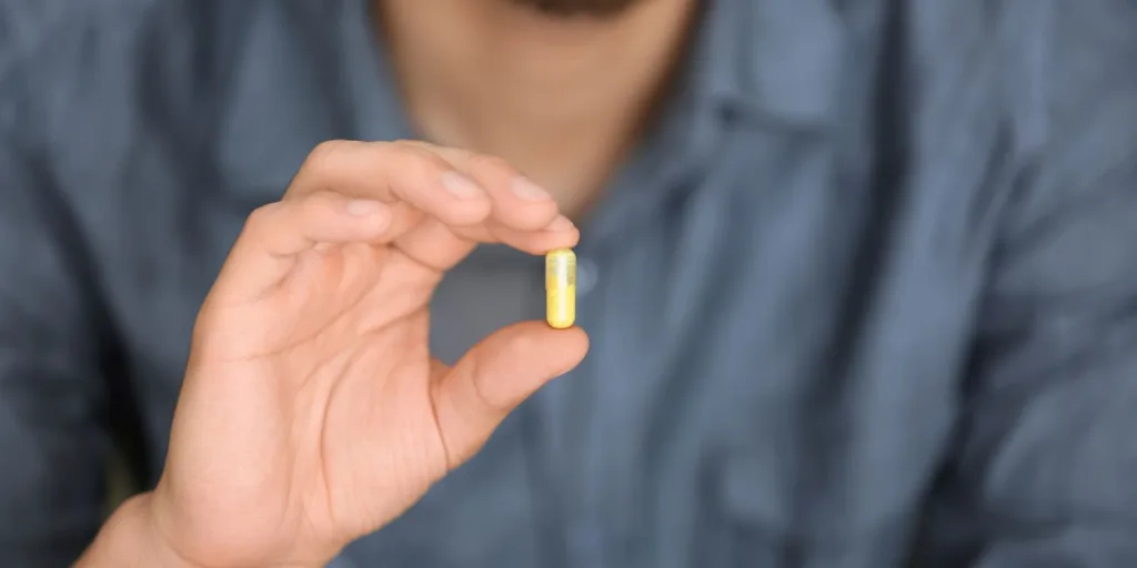 A closeup shot of a man's hand holding a capsule filled with yellow powder.
