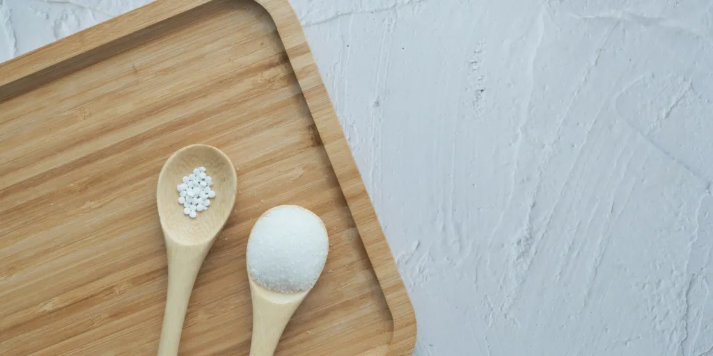 Two wooden spoons, one containing tiny sugar pills, the other containing sugar granules, against a wooden cutting board.