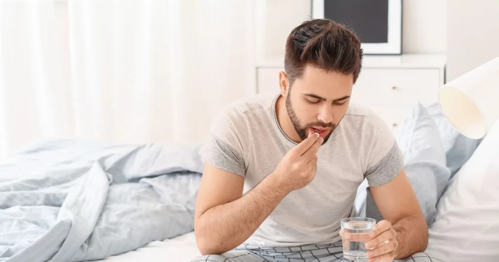 A man wearing pajamas on an unmade bed consuming a supplement and holding a glass of water.