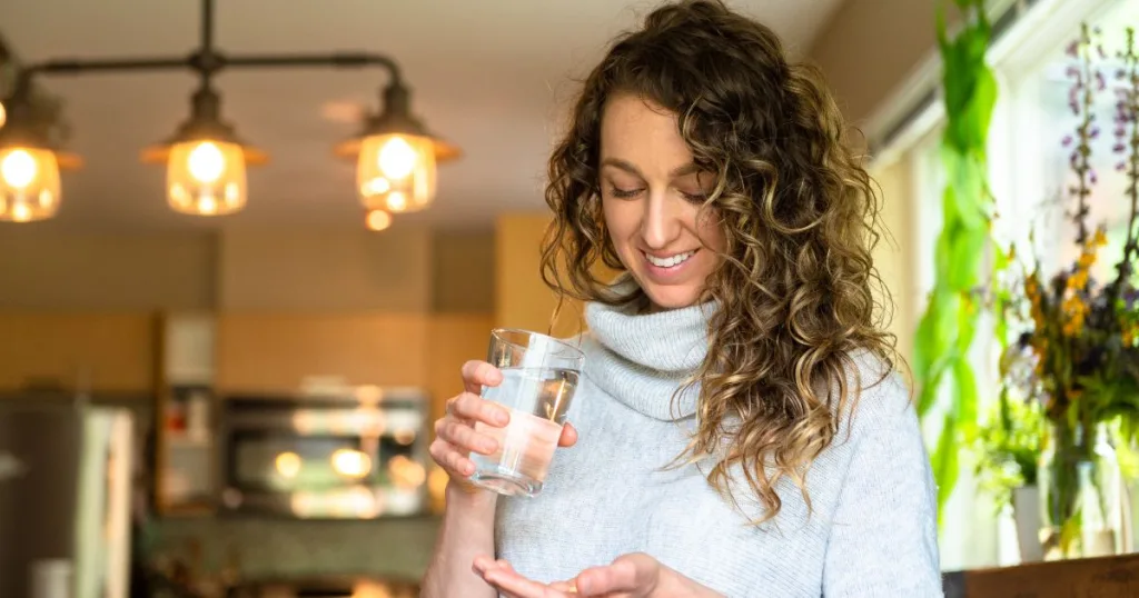 A smiling woman standing in a home kitchen with a glass of water in one hand and a supplement in the other.