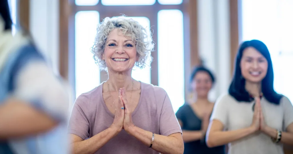 A sitting, smiling older woman in focus surrounded by others holding a namaste pose during yoga.