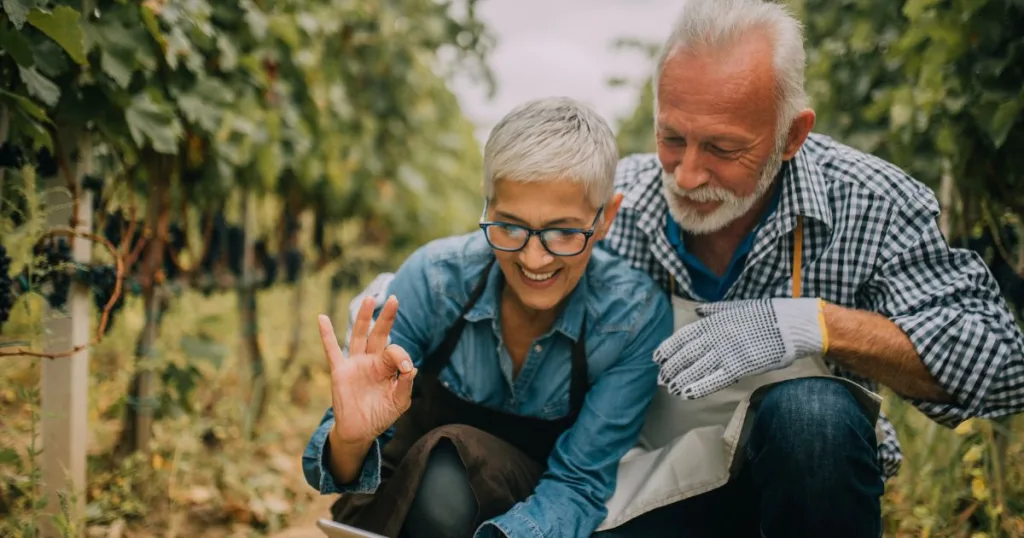 An older man and woman bending down and using a cell phone camera to take a selfie in an orchard setting.