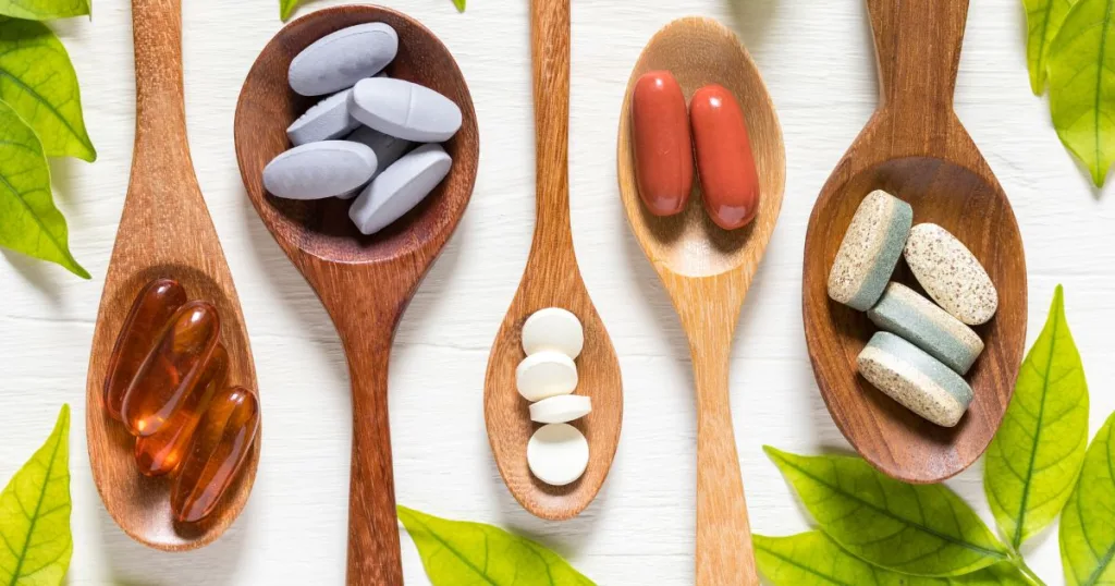 Surrounded by leaves, five spoons in alternating sizes containing supplements in various colors, shapes, and sizes.