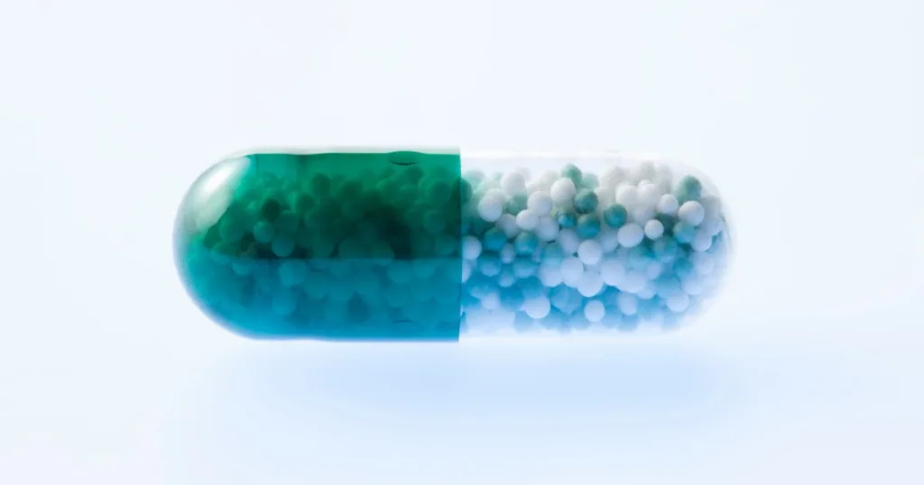 A close up shot of a see-through clear and green capsule filled with small pellets against a white background.