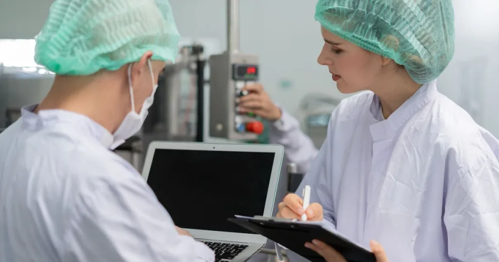 Two bulk capsule manufacturing workers in personal protective equipment inspecting results from a quality assurance machine.