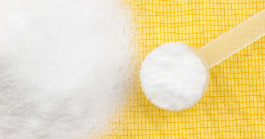 White Creatine powder in a Pile Next to a Small Scoop filled with it against a yellow background.