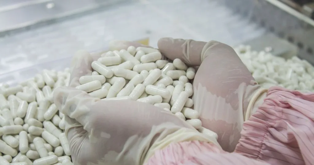 Two gloved hands picking up filled white capsules from a pile in a manufacturing environment.