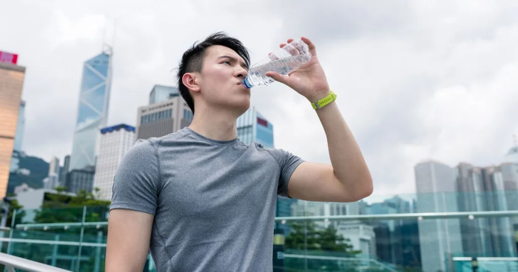 A man in workout clothes walking on a bridge in the city while drinking a bottle of water.