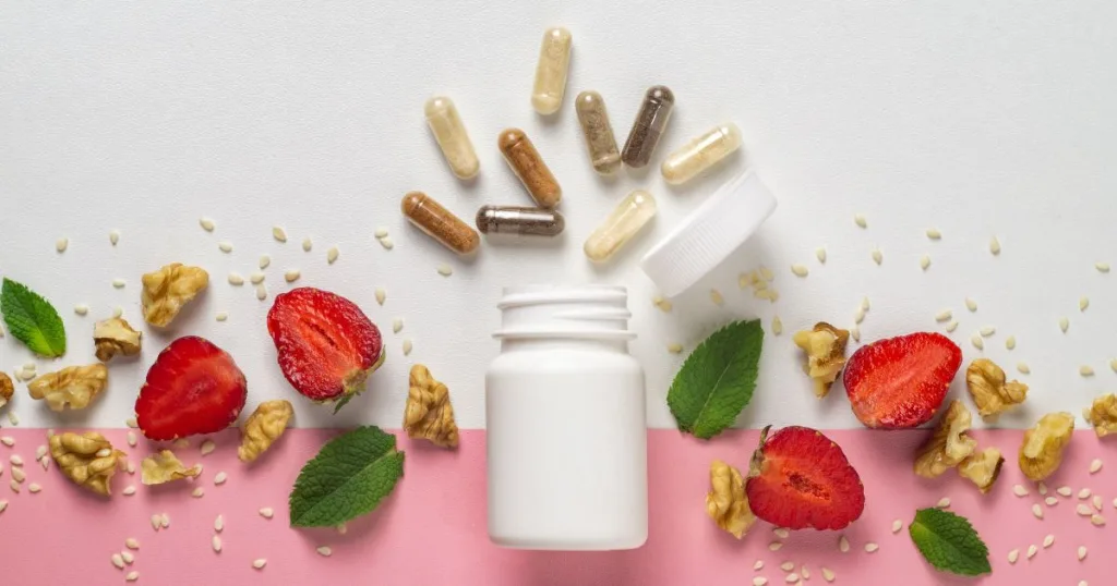 Filled supplement capsules coming out of a bottle surrounded by strawberries, mint leaves, sesame seeds, and walnuts.