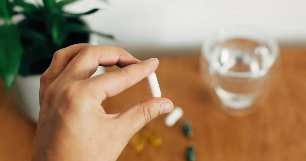 A close up of a person's hand holding a filled capsule supplement against a blurry home setting background.
