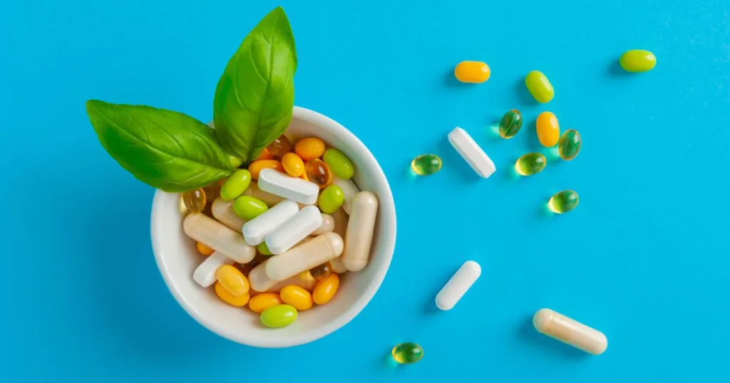 A variety of natural supplements (including capsules, tablets, and gel capsules) scattered and in a bowl with two leaves.