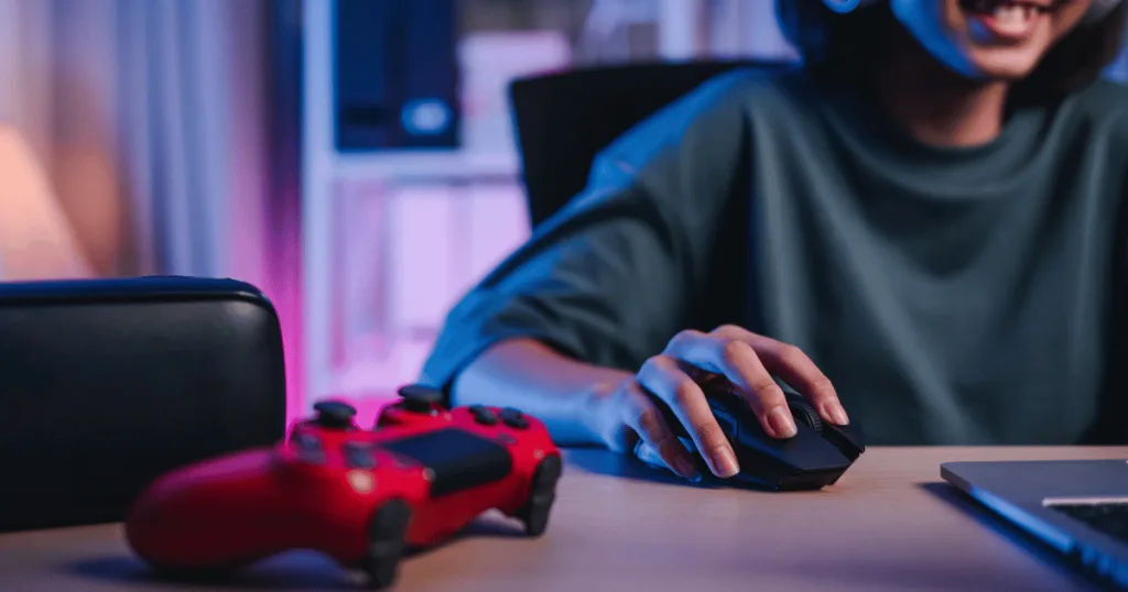 A close up of a smiling gamer with their hand on a computer mouse next to a video game controller.
