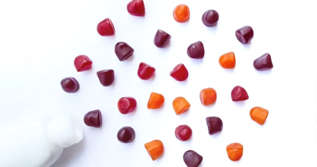 White multi-colored magnesium gummies spilling out of a white supplement bottle against a white background.
