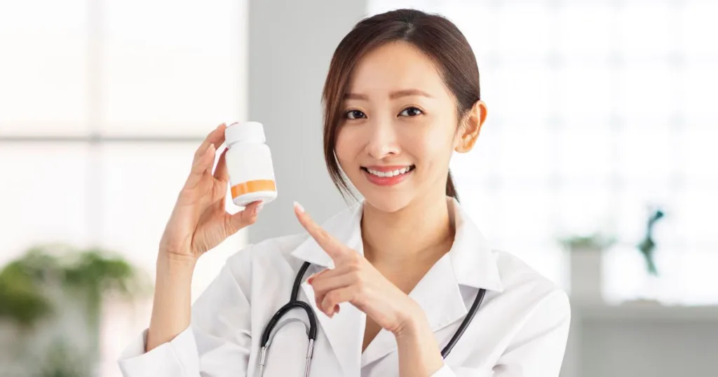 A smiling doctor wearing a white coat with a stethoscope around her neck. She is pointing to a bottle of supplements.