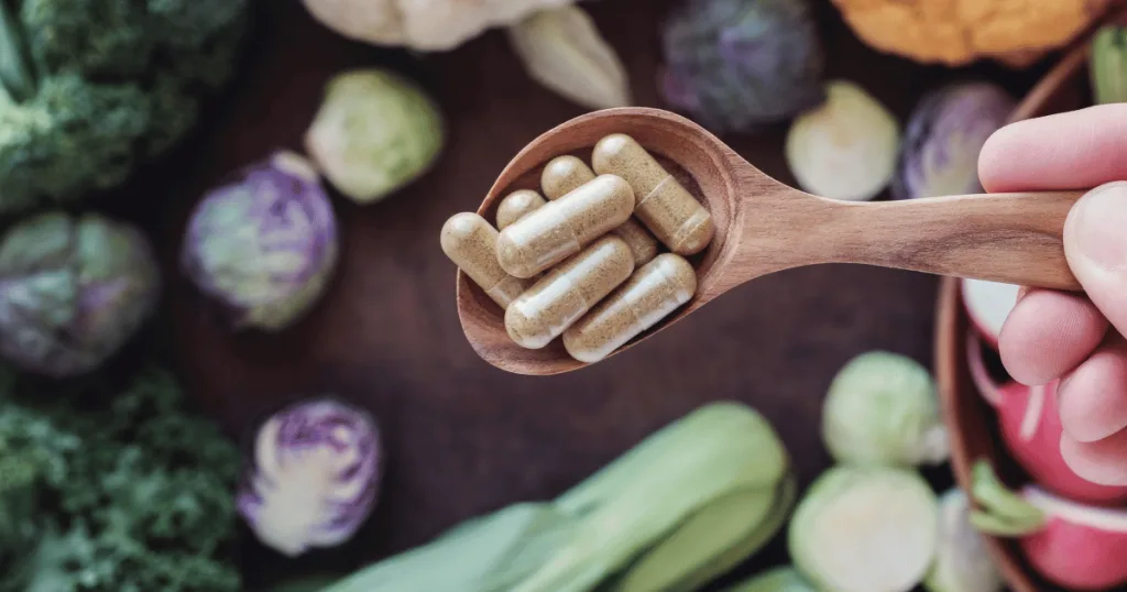 In the foreground, a hand holding a wooden spoon with filled clear capsules. In the background, various fruits and vegetables.