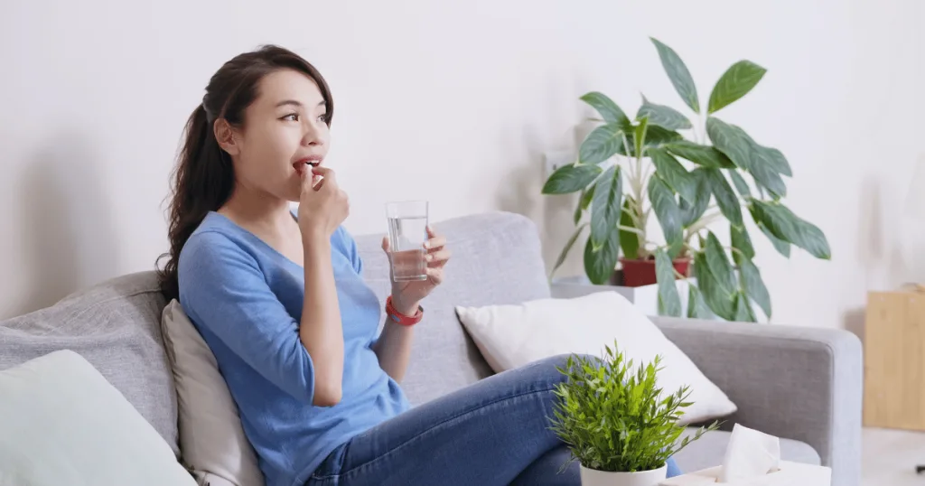 A woman sitting on a couch next two a couple of houseplants holding a glass of water and placing a magnesium supplement in her mouth.