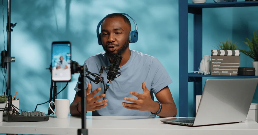 A male influencer wearing headphones talking into a mic and recording himself on a smartphone.