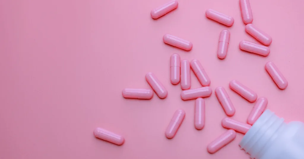 Pink capsules spilling out of a women's supplement bottle against a pink background.