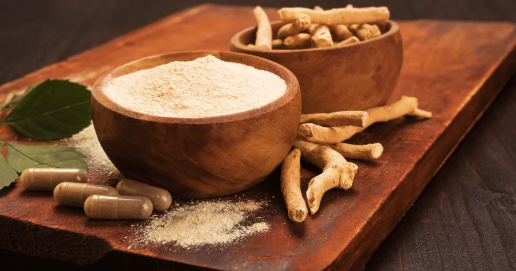 A bowl filled with powdered ashwagandha, surrounded by capsules and another bowl filled with ashwagandha roots and leaves.
