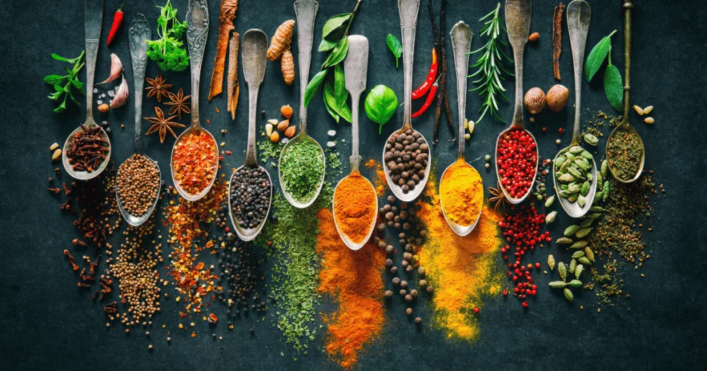 Spoons filled with various herb, such as turmeric powder, black peppercorns, ginger, star anise, nutmeg, and more.