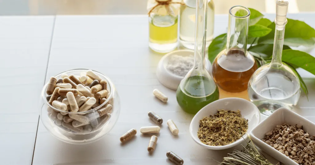 A clear glass bowl containing capsules filled with tan and brown powders. It is surrounded by different nutraceutical ingredients, liquids, oils, and powders.