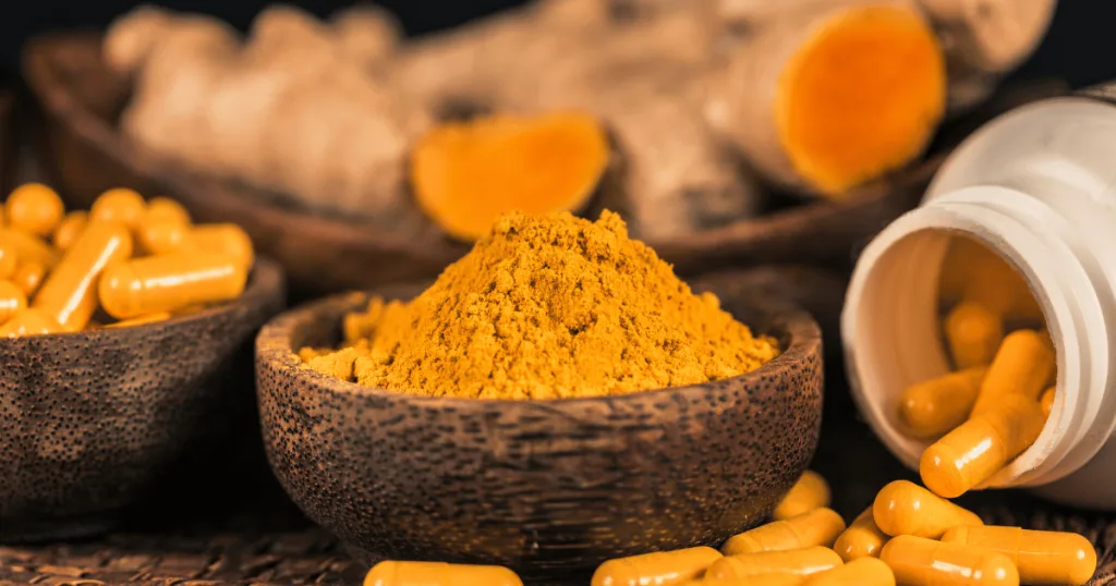 A bowl of yellow turmeric powder at the forefront, with a bowl of capsules filled with yellow powder against a background of turmeric roots.
