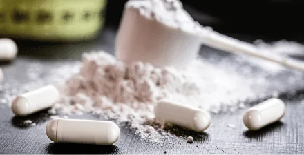 Capsules filled with white powder in the foreground, with a pile of white powder and measuring cup in the background.