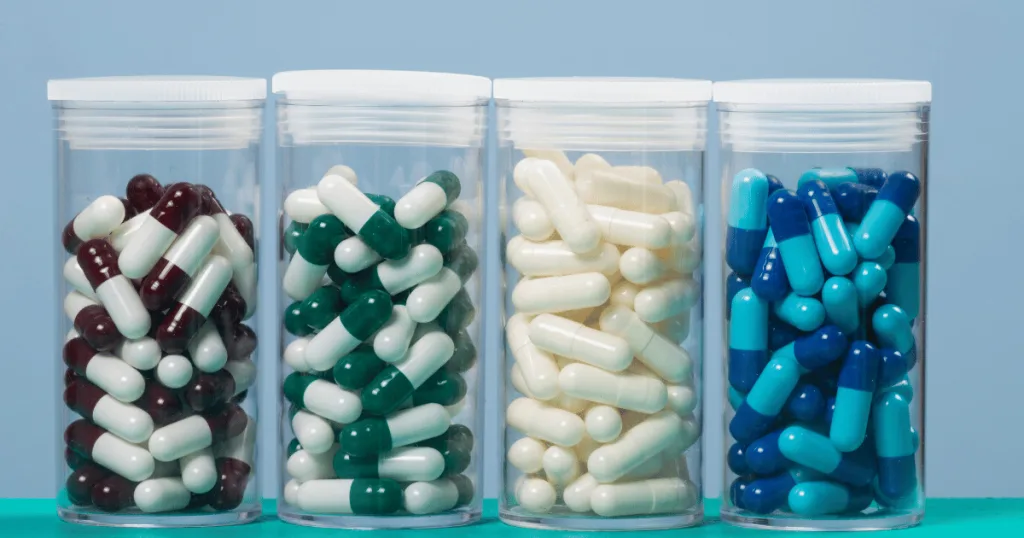 Four clear containers lined up. One is filled with maroon and white capsules, the second with green and white capsules, the third with all-white capsules, and the fourth with light blue and dark blue capsules.