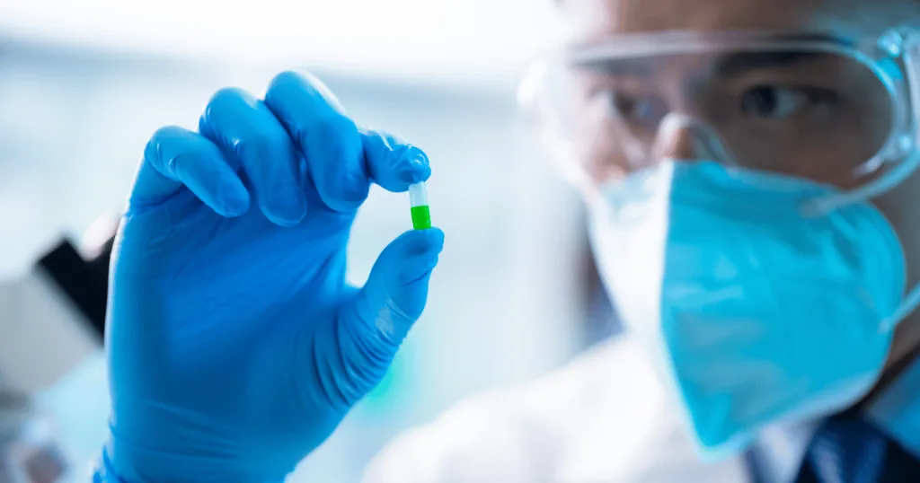 A scientist wearing PPE inspecting a green and white capsule up close, holding it between his thumb and forefinger.