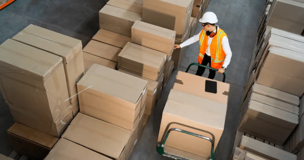 A warehouse worker pushing a dolly, surrounded by packaged products.