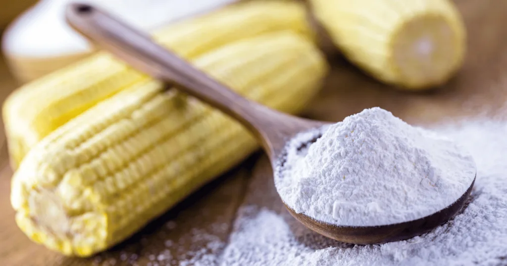 A wooden spoon filled with corn starch, with corn cobs in the background.