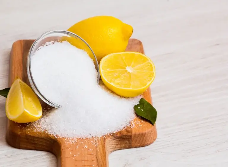 Wooden board that is holding sliced and whole lemon along with an overturned bowl with citric acid powder spilled out.