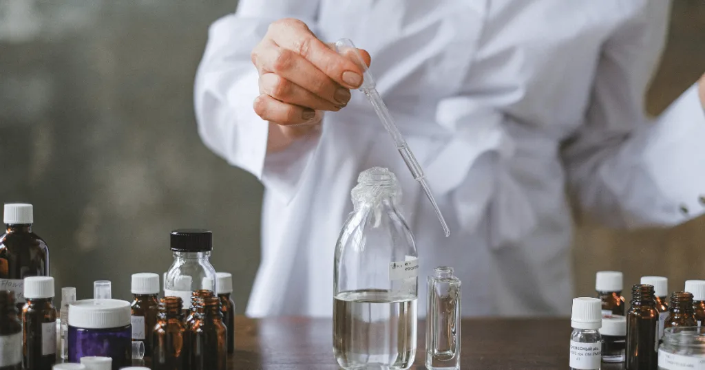 A person in a white coat filling a dropper of clear glycerin. It looks like they are testing/formulating personal care products.