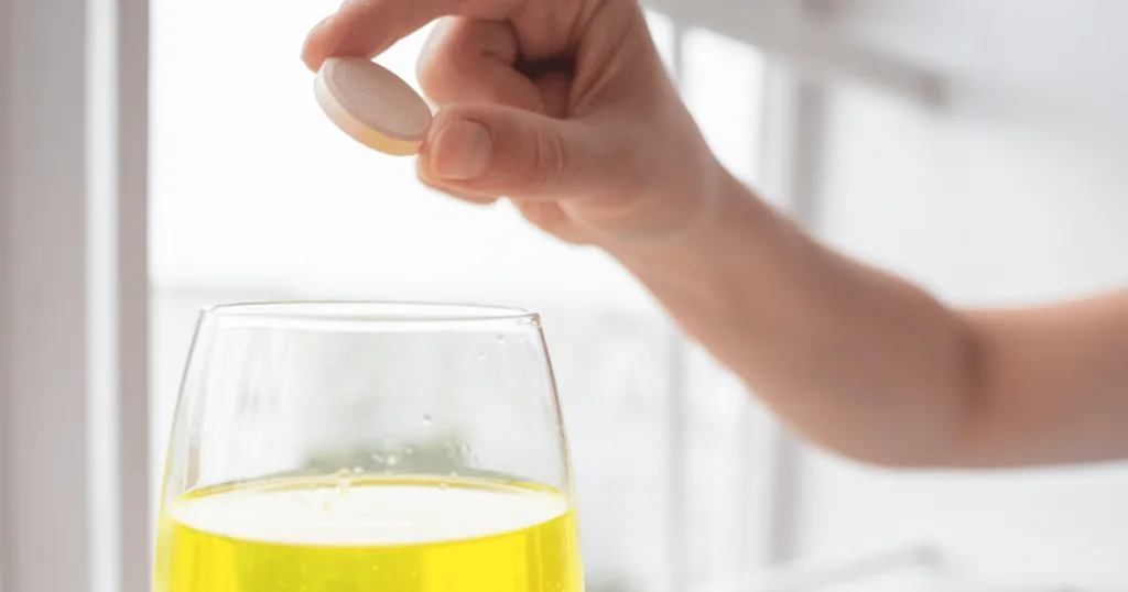 A hand holding a large vitamin c effervescent tablet over a glass filled with yellow liquid.