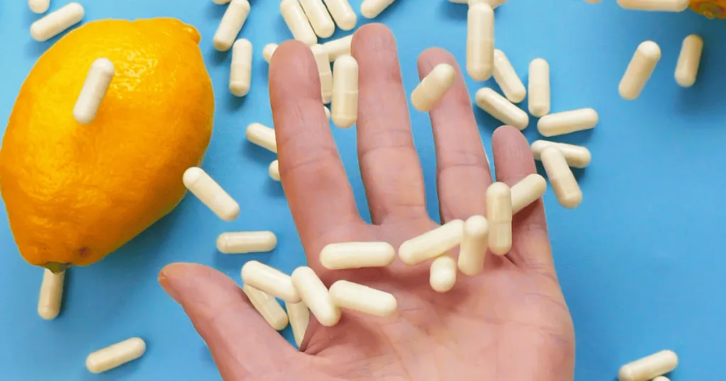 A still shot of light yellow capsules spilling into a person's open palm against a blue background. There is an orange next to the capsules on the ground.