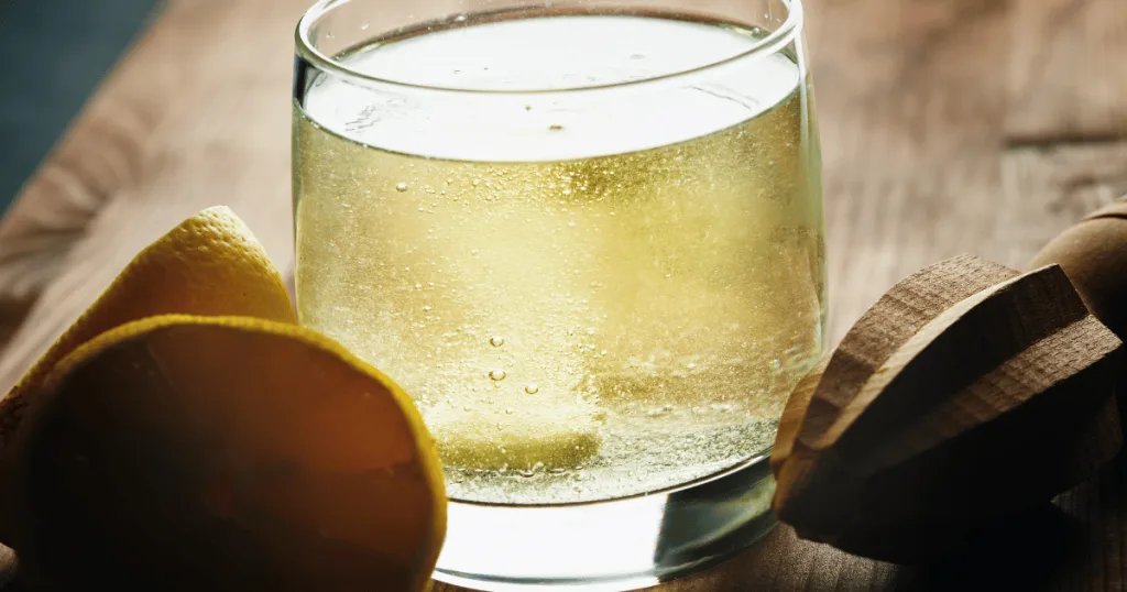 A clear class containing water and a dissolving effervescent vitamin c tablet. A couple of orange slices along with a juicer are next to the glass.