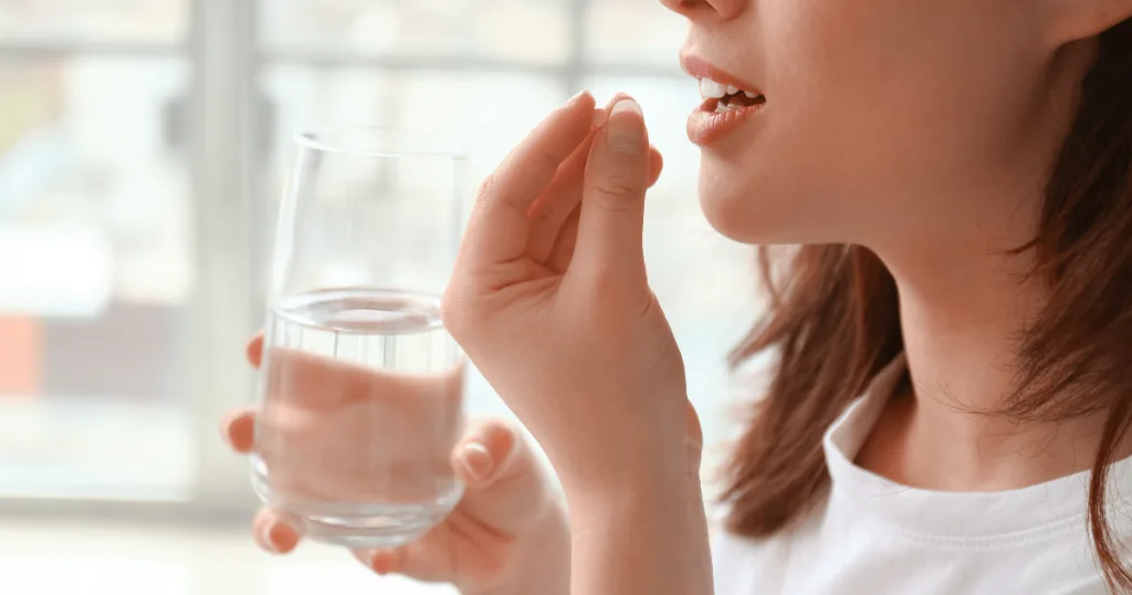 A woman holding a glass of water in one hand, holding a brain supplement in tablet format up to her mouth to consume.