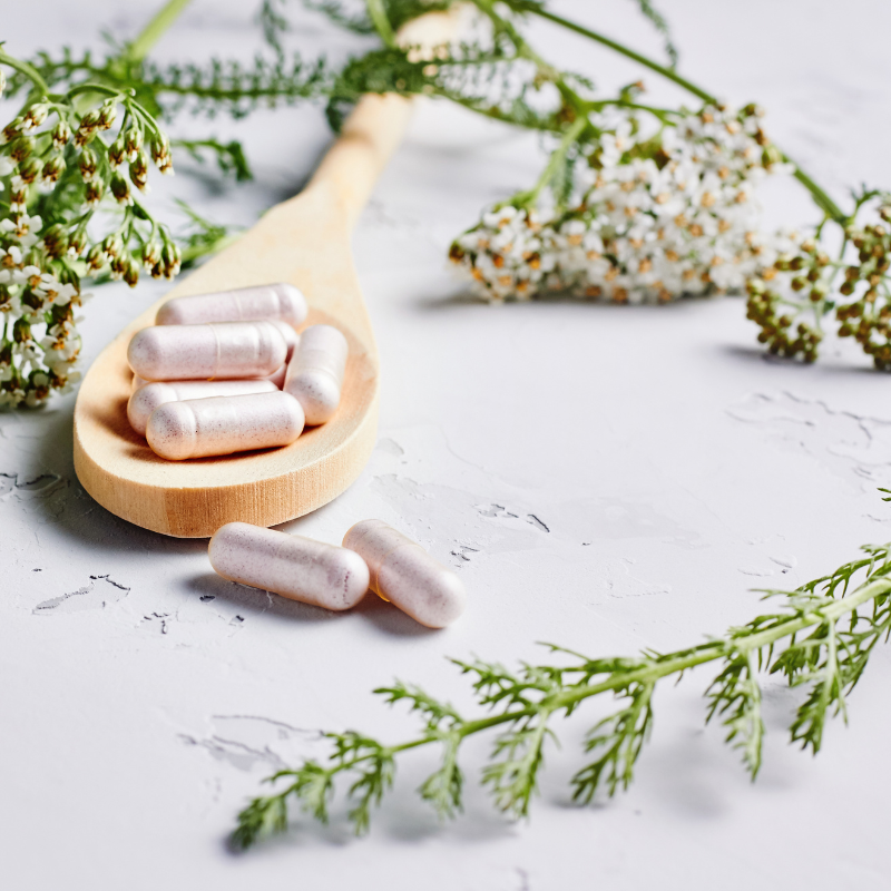 Clear capsules filled with white material in spoon surrounded by herbs.