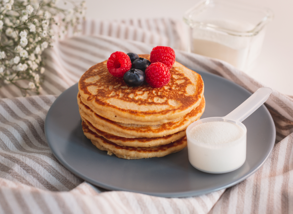 Scoop of whey protein next to stack of pancakes with raspberries and blueberries on top.