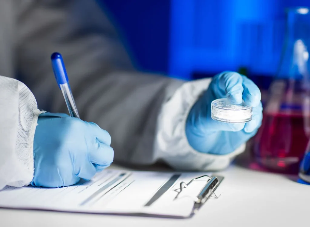 Close up of person wearing laboratory PPE holding a container of powder while writing data on a clipboard.