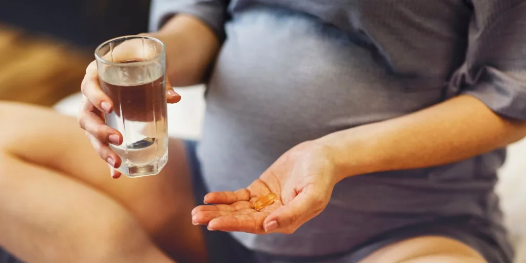 Closeup on pregnant woman's hands holding a glass of water and a gel cap supplement.